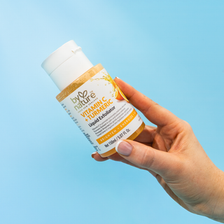 Hand holding a bottle of 'By Nature' Vitamin C + Turmeric Liquid Exfoliator against a blue background